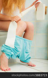 Woman with constipation or diarrhoea sitting on toilet with her blue pajamas down around her legs, holding toilet paper ready in her hands