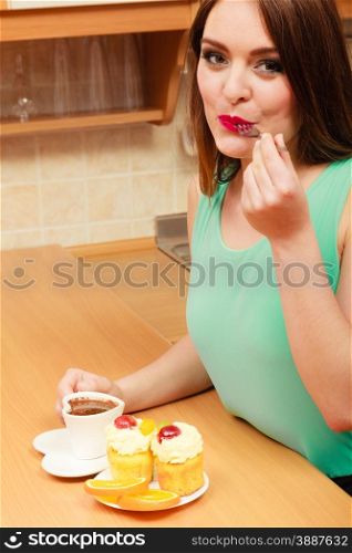 Woman with cup of coffee eating delicious gourmet sweet cream cake cupcake and orange. Glutton girl sitting in kitchen with hot beverage having breakfast. Appetite and gluttony concept.. Woman with coffee eating cream cake. Gluttony.