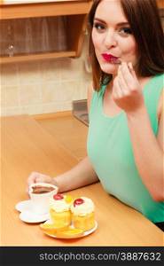 Woman with cup of coffee eating delicious gourmet sweet cream cake cupcake and orange. Glutton girl sitting in kitchen with hot beverage having breakfast. Appetite and gluttony concept.. Woman with coffee eating cream cake. Gluttony.