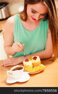 Woman with cup of coffee forking delicious gourmet sweet cream cake cupcake and orange. Glutton girl sitting in kitchen with hot beverage having breakfast. Appetite and gluttony concept.. Woman with coffee and cake in kitchen. Gluttony.