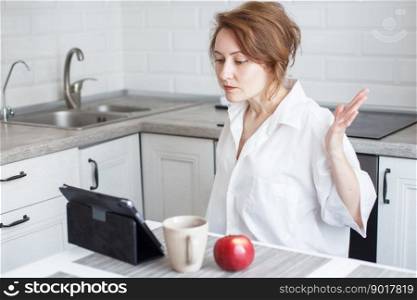 woman with cup of coffee or tea using laptop in quarantine lockdown in kitchen in white shirt. girl with a tablet. woman with cup of coffee or tea using laptop in quarantine lockdown in the kitchen in white shirt. girl with a tablet