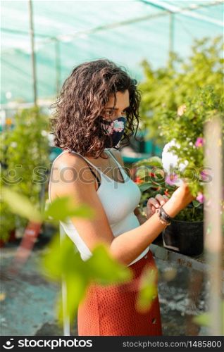 Woman with face mask gardening in greenhouse. People outdoor activities enjoying nature during pandemic coronavirus crises. Female with diy mask trought plants and flowers. Woman with face mask gardening in greenhouse covid-19
