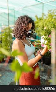 Woman with face mask gardening in greenhouse. People outdoor activities enjoying nature during pandemic coronavirus crises. Female with diy mask trought plants and flowers. Woman with face mask gardening in greenhouse covid-19