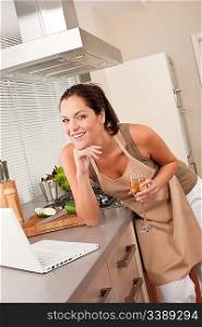 Woman with glass of white wine and laptop in the kitchen cooking