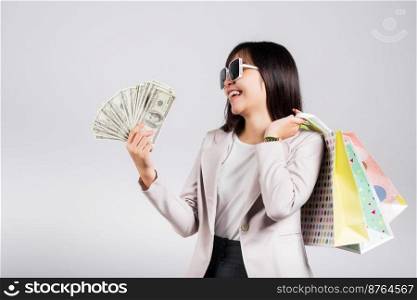 Woman with glasses confident shopper smile holding online shopping bags multicolor and dollar money banknotes on hand, excited happy Asian young female person studio shot isolated on white background