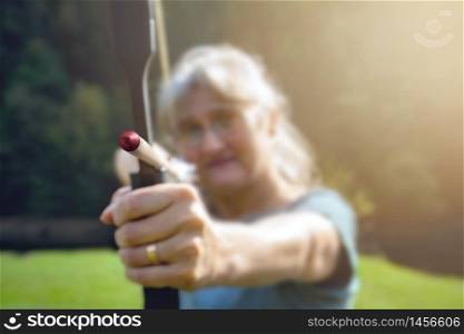 Woman with gray hair aiming with bow and arrow in nature and sun. Concept for the fight to reach the target