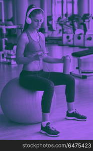 woman with headphones in fitness gym. young healthy woman drinking water in fitness gym while sitting on pilates ball and listening music on headphones from smartphone duo tone