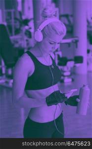 woman with headphones in fitness gym. young healthy woman drinking water in fitness gym while sitting on pilates ball and listening music on headphones from smartphone duo tone