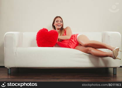 Woman with heart shape pillow. Valentines day love. Smiling pretty cute young woman girl with red heart shape pillow sitting on white sofa couch. Valentines day love.