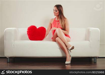 Woman with heart shape pillow. Valentines day love. Smiling pretty cute young woman girl with red heart shape pillow sitting on white sofa couch. Valentines day love.