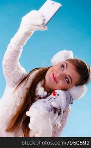 Woman with little snowman taking selfie photo.. Pretty cute woman holding little snowman taking selfie self photo picture with camera. Attractive girl in earmuffs and white sweater pullover in studio on blue. Winter fashion.