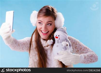 Woman with little snowman taking selfie photo.. Smiling pretty cute woman holding little snowman taking selfie self photo picture with camera. Attractive girl in earmuffs and white sweater pullover in studio on blue. Winter fashion.