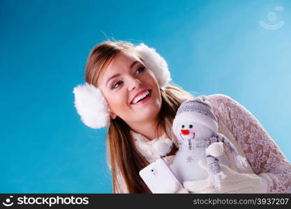 Woman with little snowman taking selfie photo.. Smiling pretty cute woman holding little snowman taking selfie self photo picture with camera. Attractive girl in earmuffs and white sweater pullover in studio on blue. Winter fashion.