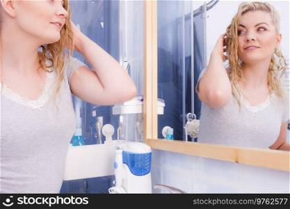 Woman with long wet curly hair in bathroom looking in mirror. Blonde girl taking care refreshing her hairstyle in morning. Haircare concept.. Woman looking at herself in bathroom mirror