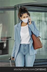 woman with medical mask luggage talking smartphone airport during pandemic