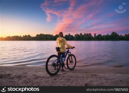 Woman with mountain bike on the river coast at sunset in summer. Landscape with young girl, bicycle, sandy beach, purple sky with colorful pink clouds, water at dusk. Sport and travel. Cycle. Nature