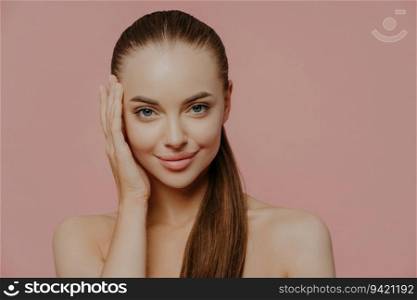 Woman with ponytail enjoys fresh skin, charming look, bare shoulders, isolated on pink wall. Health care, makeup, spa.