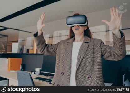 Woman with surprised facial expression experiencing amazing world of virtual reality with VR goggles, standing alone in front of office desk, holding her hands in air, using 3d glasses at work. Surprised woman experiencing amazing world of virtual reality with VR goggles at work