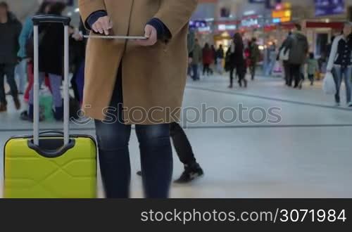 Woman with tablet PC in hands standing in airport or railway station building, her trolley bag is standing by her.