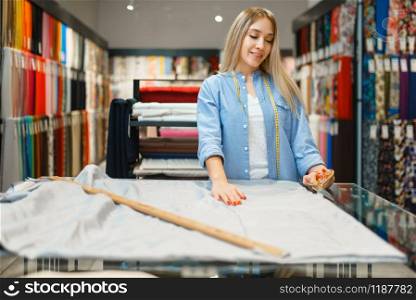 Woman with tape measures the fabric closeup in textile store. Shelf with cloth for sewing on background, clothing patterns choice in shop. Woman measures the fabric closeup, textile store