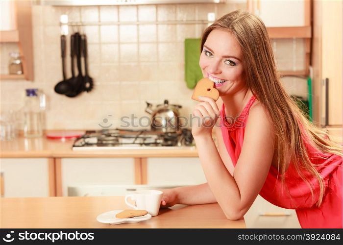 Woman with tea coffee eating gingerbread cookie.. Happy woman with tea or coffee eating heart shape gingerbread cookie. Pretty gorgeous young girl with hot beverage relaxing in kitchen.