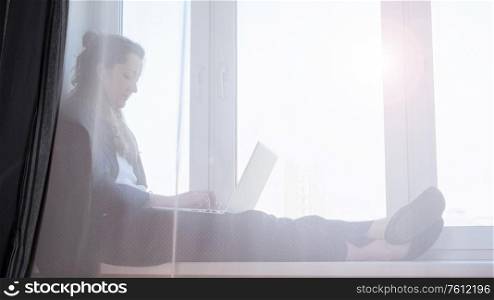 Woman working on laptop from home due to coronavirus self isolation. Sitting on window sill. Authentic home workplace. Coronavirus outbreak 2020.. Woman working from home