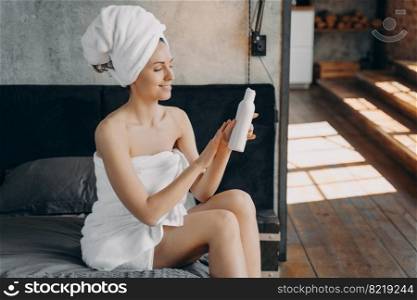 Woman wrapped in towels holding bottle of sunscreen lotion applying cosmetics after shower sitting on bed in hotel. Smiling female moisturizing skin, enjoying skincare routine on vacation. Body care. Girl wrapped in towels uses sunscreen lotion after shower sitting on bed in hotel. Body skin care