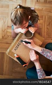 Woman writing an address on a cardboard box parcel in room at home. Little girl helping her mother to wrap a package. Preparing parcel to send