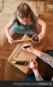 Woman writing an address on a cardboard box parcel in room at home. Little girl helping her mother to wrap a package. Preparing parcel to send