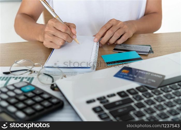 Woman writing on notebook with computer, calculator and credit card on desk, account and saving concept.