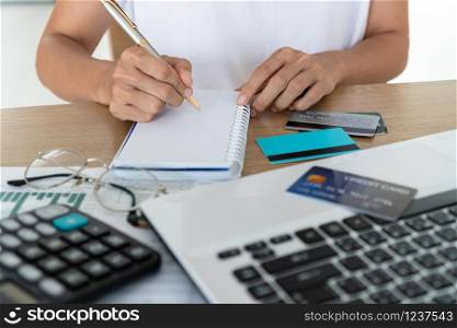 Woman writing on notebook with computer, calculator and credit card on desk, account and saving concept.