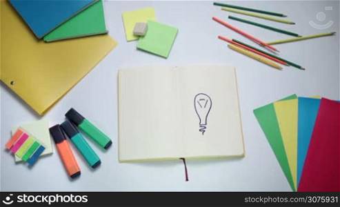 Woman writing word idea on open note pad with marker above light bulb drawing. Background white table with office accessories
