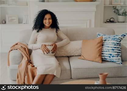 Women and maternity concept. Portrait of lovely sweet expectant mixed race lady sitting on soft couch decorated with cushions with her hands folded on belly in form of heart. Happiness of pregnancy. African future mom with bright smile on face keeping hands folded on belly in form of heart