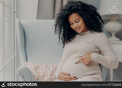 Women and pregnancy concept. Beautiful happy pregnant woman in white dress with curly hair sitting on comfortable armchair at home near window and pointing at her future baby in her belly. Future african american mother in white dress talking with baby inside belly