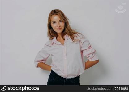 Women beauty. Elegant attractive female model in casual shirt and jeans standing with folded hands behind her back and looking at camera with flirting smile, isolated over grey studio background. Elegant attractive female model in casual shirt and jeans standing with folded hands behind her back