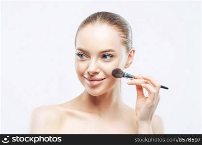 Women, cosmetology concept. Tender healthy European woman gives makeup lesson, applies cosmetic with beauty brush, looks aside with smile, isolated on white background, shows perfect naked body