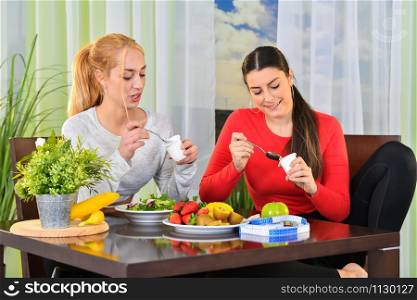 Women eating yogurt and having a conversation sitting at a table with a salad, assorted fruit and a measuring tape. Healthy lifestyle concept.. Women having a chat over a healthy meal