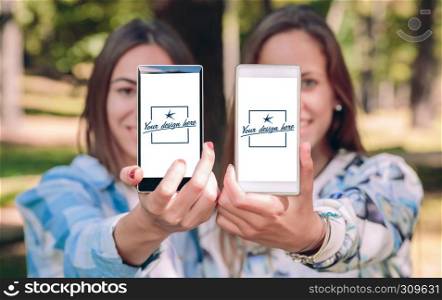 Women friends showing smartphones with their selfie photos taked over a forest background. Customizable mobile screens. Women showing smartphones with their selfie photos