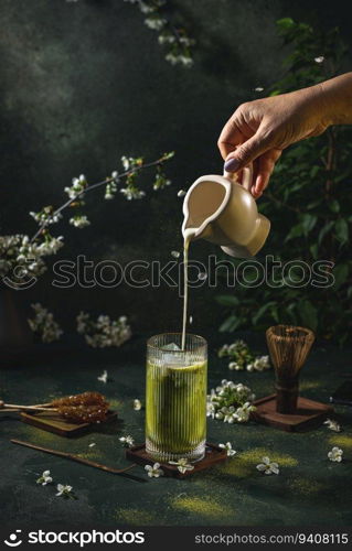 Women hand is pouring homemade sour cream from small jar to glass with matcha tea. Dark background with spring cherry blossom and traditional dishes for this japanese tea