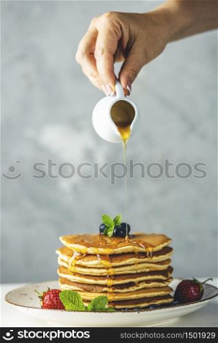 Women hand is pouring syrup from small ceramic jar to pancake with srtawberry, blueberry and mint in ceramic dish on a light wooden table and gray background.