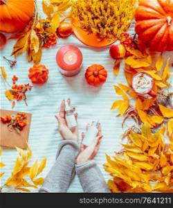 Women hands holding bottles of cosmetic products on white blanket with pumpkins, candles and fall leaves. Autumn beauty concept. Seasonal skin care . Flat lay. Top view