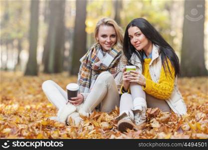 Women in autumn park drink coffee. Two cheerful female friends drinking coffee in autumn park