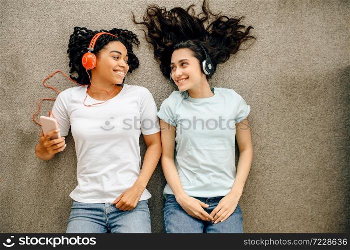 Women in headphones lying on the floor, top view. Pretty girlfriends in earphones relax in the room, music lovers resting. Women in headphones lying on the floor, top view