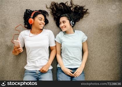 Women in headphones lying on the floor, top view. Pretty girlfriends in earphones relax in the room, music lovers resting. Women in headphones lying on the floor, top view