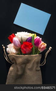 Women international day card with a bouquet of diverse flowers wrapped in a hessian bag and an unwritten blue message card on a black background.