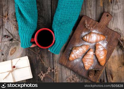 women's hands in a green knitted sweater holding a red ceramic mug with black coffee, next to baked croissants sprinkled with powdered sugar, gray wooden background