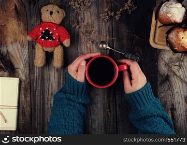 women's hands in a green knitted sweater holding a red ceramic mug with black coffee, gray wooden table, top view