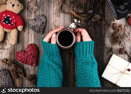 women's hands in a green knitted sweater holding a red ceramic mug with black coffee, gray wooden table