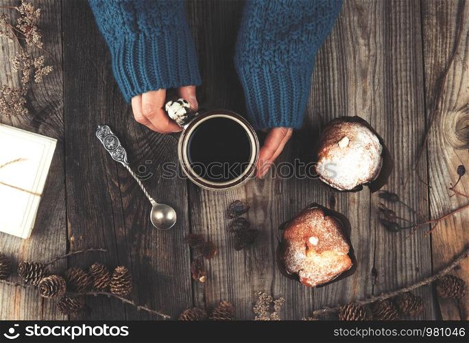 women's hands in a knitted blue sweater holding a ceramic mug with black coffee, gray wooden table, top view