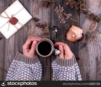 women's hands in a knitted brown sweater holding a ceramic mug with black coffee, gray wooden table, top view